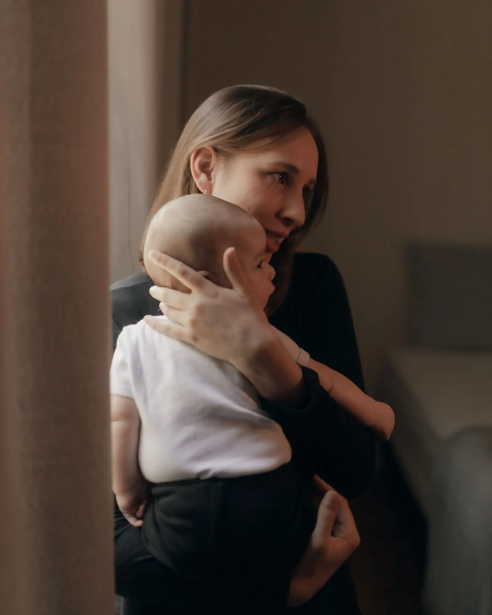 A parent holding their baby beside a sunlit window at home